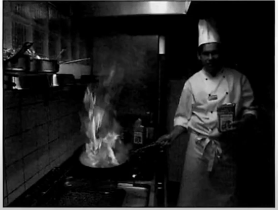 A chef standing beside a gas hob where a pan has caught alight in a restaurant kitchen