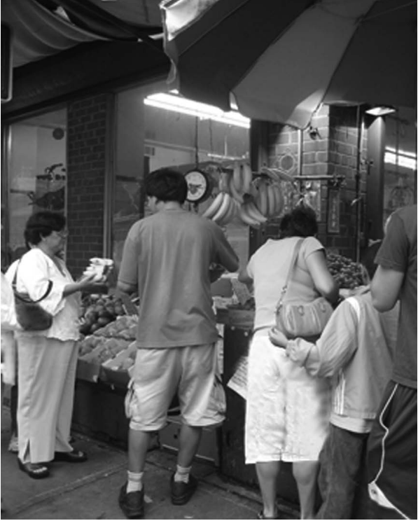 Shoppers at an outdoor fruit and vegetable stall under a large umbrella